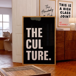 May include: A framed poster with the words "THE CULTURE." in white on a black background. Two other framed posters are visible, one with "The Homebody Club" and another with "THIS IS A HIGH CLASS JOINT." The room has wooden flooring and a wooden cabinet.