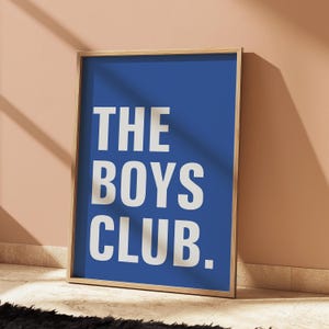 May include: A framed print with the text "THE BOYS CLUB" in large white capital letters on a blue background. The frame is a light wood color, and the print is leaning against a peach-colored wall.