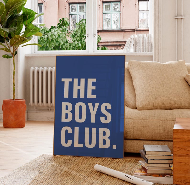 May include: A blue rectangular sign with the words 'THE BOYS CLUB.' in large white block letters. The sign is leaning against a beige couch in front of a window. A potted plant is to the left of the sign, and a stack of books is on the floor.