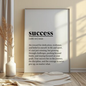 May include: A framed print with the word "success" in bold black letters at the top. Below, the definition of success is printed in black text. The print is displayed on a wooden floor, with a vase and decorative plant to the left.