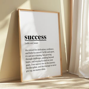 May include: A framed print with the word "success" in bold black font. The definition of success is written below the word. The frame is a light brown color, and the background is a neutral beige.
