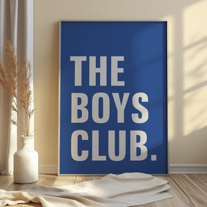 May include: A framed poster with the text "THE BOYS CLUB" in large, white, sans-serif letters against a solid blue background. The poster is displayed on a wooden floor, with a white vase and dried plants visible.