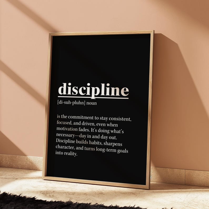 May include: A framed black and white print with the word "discipline" in a bold, sans-serif font. The definition of discipline is written below, describing commitment, focus, and achieving goals. The frame is a light brown color.