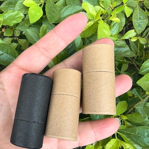 Puede incluir: Tres tubos cilíndricos de cartón, uno negro y dos marrones naturales, se sostienen en una mano sobre un fondo de follaje verde. Los tubos son de tamaño y forma similares, con una tapa extraíble.
