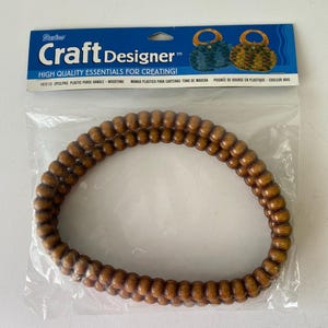 May include: A package containing a woodtone plastic purse handle. The handle is made of small, rounded beads strung together in a semi-circular shape. The package has the text "Craft Designer" and "High Quality Essentials for Creating!"