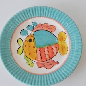 Puede incluir: Un plato redondo de cerámica con un diseño de pez pintado a mano. El pez es amarillo, naranja y azul, con detalles en verde y rosa. El plato tiene un borde turquesa con estrías verticales y un centro blanco.