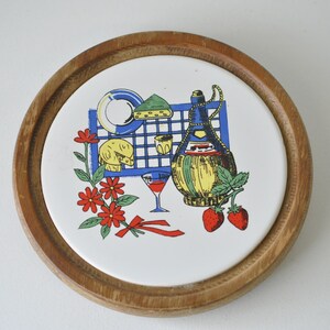 May include: A round wooden coaster with a white ceramic tile insert. The tile features a colorful illustration of a wine bottle, a glass of wine, cheese, and strawberries. The design is reminiscent of a picnic or Italian countryside.