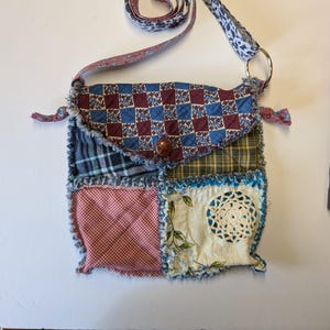May include: A patchwork crossbody bag featuring a flap with a blue and maroon checkered floral pattern, secured by a brown button. The body consists of quilted squares in plaid, gingham, and cream fabric with a circular lace appliqué and frayed edges.