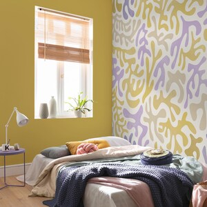 May include: A bedroom with a yellow wall and a bed with a purple, yellow and white patterned headboard. The bed has a grey blanket and a pink throw pillow. There is a small table next to the bed with a lamp and a book.