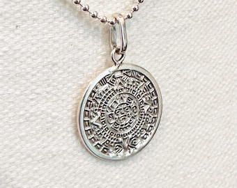 May include: A silver-toned circular pendant featuring an intricate embossed Aztec calendar design, suspended from a matching silver-toned ball chain necklace. The pendant has a polished metallic finish with detailed carvings of symbols and a central face.