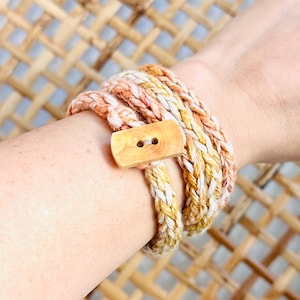 May include: A multi-colored braided cord bracelet with a wooden button closure. The bracelet is made with shades of peach, white, and yellow.