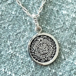 May include: A silver-colored pendant necklace featuring a circular pendant with an intricate, detailed design. The pendant is suspended from a delicate chain. The pendant has the text "925" on the bail.