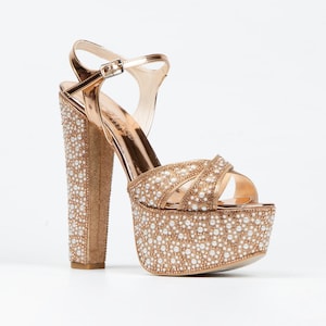 Puede incluir: Sandalias de plataforma doradas con un tacón grueso y una correa alrededor del tobillo. Las sandalias están cubiertas de pequeños diamantes de imitación blancos.