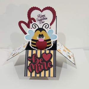 May include: A pop-up Valentine's Day card featuring a cartoon bee with blue wings and a red heart. The card has a heart-shaped message that reads "Happy Valentine's Day" and a box that says "Be Mine". The box has black and yellow stripes.