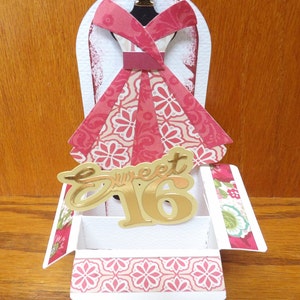 May include: A pink and white paper dress with a gold 'Sweet 16' cutout on top of a white box with a pink and green patterned design.