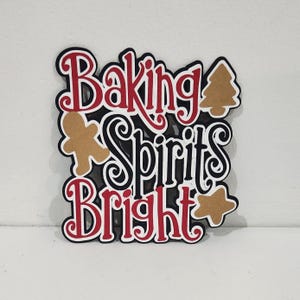 May include: A layered paper cutout with the words "Baking Spirits Bright" in red and black, with gingerbread man, star, and Christmas tree shapes. The design is festive and holiday-themed.