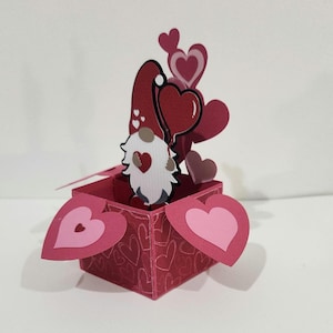 May include: A red pop-up box with a gnome holding a heart-shaped balloon. The box is decorated with heart patterns and has heart-shaped flaps. Several heart cutouts are arranged around the gnome, creating a Valentine's Day theme.