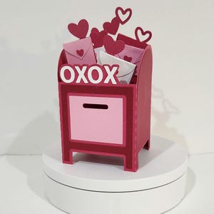 May include: A Valentine's Day-themed mailbox in shades of red and pink, filled with heart-shaped cutouts and envelopes. The word "OXOX" is displayed on the front. The mailbox is on a white surface.
