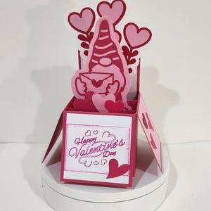 May include: A pink Valentine's Day pop-up card featuring a gnome holding an envelope and hearts. The card has the words "Happy Valentine's Day" on the front. The card is displayed on a white surface.