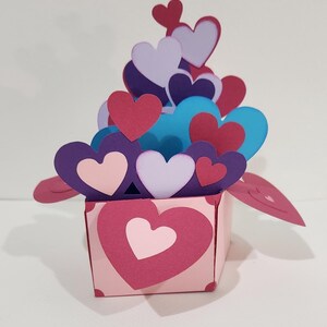 May include: A pink paper box with a large heart on the front, filled with layered paper hearts in shades of red, purple, blue, and white. The hearts are of varying sizes, creating a 3D effect.