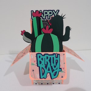 May include: A pop-up card featuring a cactus design. The card is open, revealing a green cactus with pink flowers and the words "Happy Birthday" on the front.