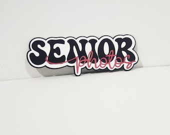 May include: A decorative sign with the words "SENIOR photos" in a retro font. The word "SENIOR" is in a dark blue colour with a white outline, and the word "photos" is in a pink script font.