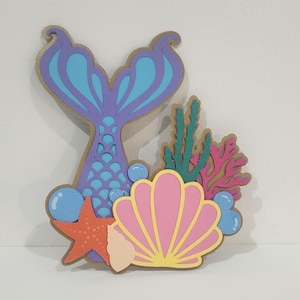 May include: A layered wooden cutout featuring a purple and blue mermaid tail, a pink seashell, an orange starfish, green seaweed, and light blue bubbles. The design is composed of multiple stacked pieces with distinct colors and textures.