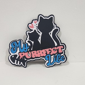 May include: A black cat silhouette with a smaller cat and pink hearts. The phrase "My Purrfect Life" is written in blue and pink, with a fish bone graphic. The design is on a white background.