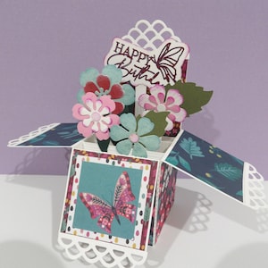 May include: A pop-up birthday card featuring a butterfly and floral design. The card is decorated with pink, blue, and white flowers, and a sign that reads "Happy Birthday". The card has a decorative, scalloped edge.