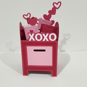 May include: A Valentine's Day-themed mailbox in shades of pink and red. The mailbox is decorated with the letters "XOXO" and contains paper hearts and envelopes. Several heart-shaped cutouts are also visible.