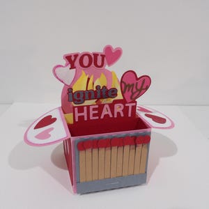 May include: A handmade Valentine's Day pop-up card with a red box design. The card features the words "YOU ignite my HEART" with heart and flame accents. The box is decorated with matchstick-like elements.