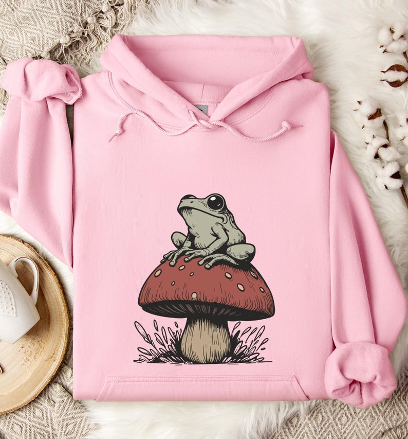 May include: A pink hoodie with a graphic of a green frog sitting on a red and white mushroom.
