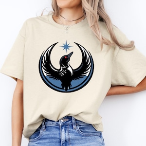 Puede incluir: Camiseta beige con un dise&ntilde;o de p&aacute;jaro negro con las alas extendidas, un c&iacute;rculo azul y una estrella. El p&aacute;jaro tiene un ojo rojo. El dise&ntilde;o est&aacute; centrado en la parte delantera de la camiseta.