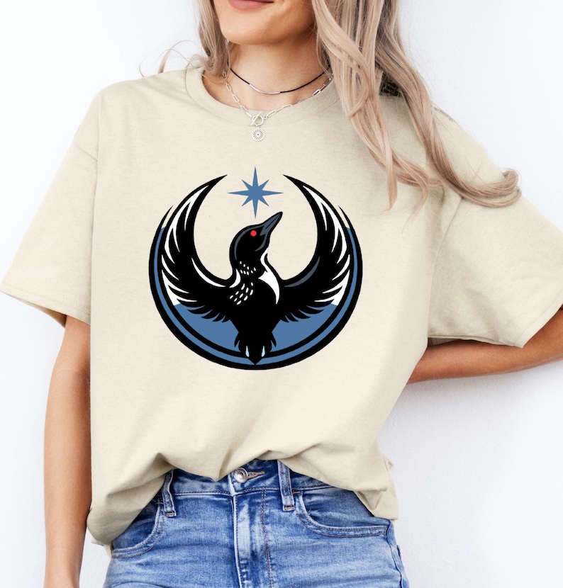 Puede incluir: Camiseta beige con un dise&ntilde;o de p&aacute;jaro negro con las alas extendidas, un c&iacute;rculo azul y una estrella. El p&aacute;jaro tiene un ojo rojo. El dise&ntilde;o est&aacute; centrado en la parte delantera de la camiseta.