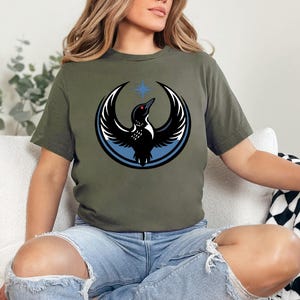 Puede incluir: Camiseta verde oliva con un gr&aacute;fico de un p&aacute;jaro negro con las alas extendidas, un ojo rojo y una estrella azul encima. El p&aacute;jaro est&aacute; rodeado por un anillo azul y detalles en blanco. La camiseta est&aacute; hecha de un material suave.