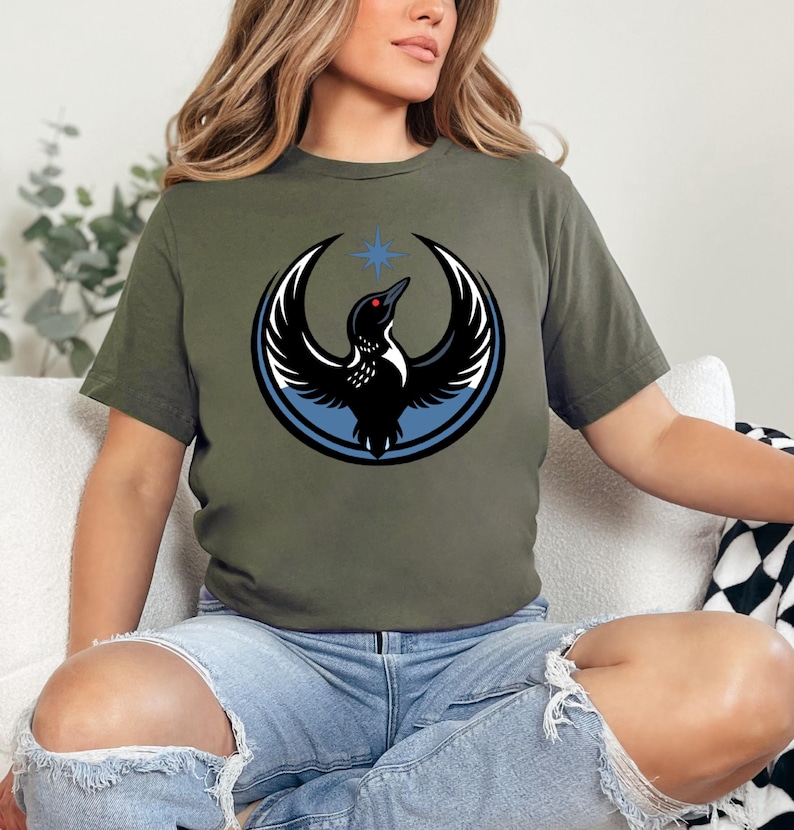 Puede incluir: Camiseta verde oliva con un gr&aacute;fico de un p&aacute;jaro negro con las alas extendidas, un ojo rojo y una estrella azul encima. El p&aacute;jaro est&aacute; rodeado por un anillo azul y detalles en blanco. La camiseta est&aacute; hecha de un material suave.