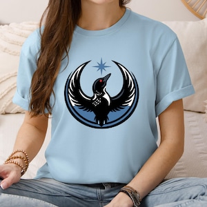Puede incluir: Camiseta azul claro con un dise&ntilde;o gr&aacute;fico de un p&aacute;jaro negro con las alas extendidas, un ojo rojo y una estrella encima. El p&aacute;jaro est&aacute; rodeado por un c&iacute;rculo azul. La camiseta es de material suave.