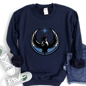 May include: Navy blue sweatshirt featuring a circular graphic of a bird with outstretched wings, a star, and a blue outline. The bird is black and white with a red eye. The sweatshirt is a comfortable, casual style.