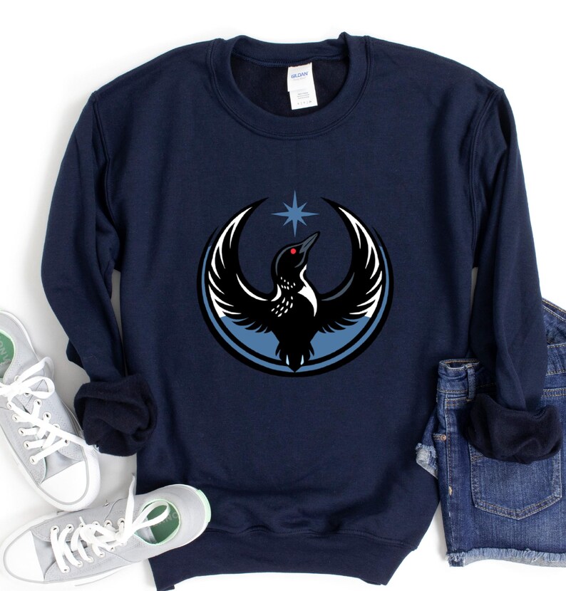 May include: Navy blue sweatshirt featuring a circular graphic of a bird with outstretched wings, a star, and a blue outline. The bird is black and white with a red eye. The sweatshirt is a comfortable, casual style.