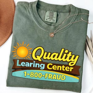 May include: Olive green Comfort Colors t-shirt with a graphic that reads "Quality Learing Center 1-800-FRAUD" in yellow, brown, and blue. A gold necklace with a small pendant rests on the shirt. A brown hat is visible in the upper left corner.