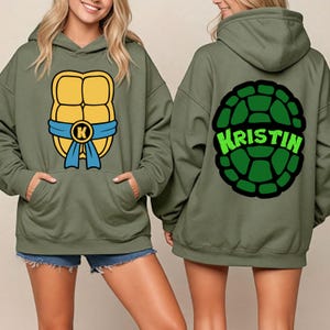 May include: Olive green hooded sweatshirt featuring a cartoon turtle shell design on the front and back. The front design includes a yellow shell, blue sash, and a letter "K". The back of the sweatshirt has a green shell design with the name "KRISTIN" in green.