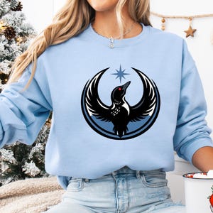 May include: Light blue sweatshirt featuring a graphic of a black bird with a red eye, wings spread, and a star above. The bird is inside a blue and black circle. The sweatshirt is a solid light blue color.