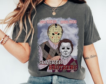 May include: Dark gray t-shirt featuring a graphic of Jason Voorhees and Michael Myers with the text "Slasher Brothers." The design includes a masked figure and a figure with a white mask, set against a cloudy background. The shirt is a casual wear item.