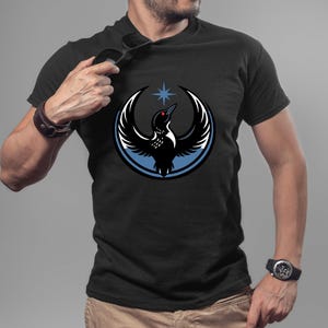 Puede incluir: Camiseta negra con un dise&ntilde;o estilizado de un ave dentro de un c&iacute;rculo azul y blanco, con una estrella encima. El ave tiene un ojo rojo y alas extendidas. La camiseta es de manga corta.