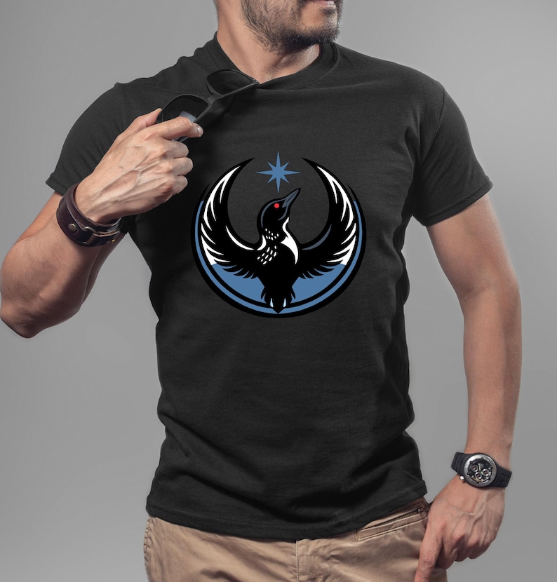 Puede incluir: Camiseta negra con un dise&ntilde;o estilizado de un ave dentro de un c&iacute;rculo azul y blanco, con una estrella encima. El ave tiene un ojo rojo y alas extendidas. La camiseta es de manga corta.