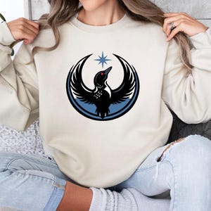May include: Cream-colored sweatshirt featuring a black bird design within a blue circle, with a star above. The bird has a red eye and is set against a cream background. The sweatshirt is a casual style.