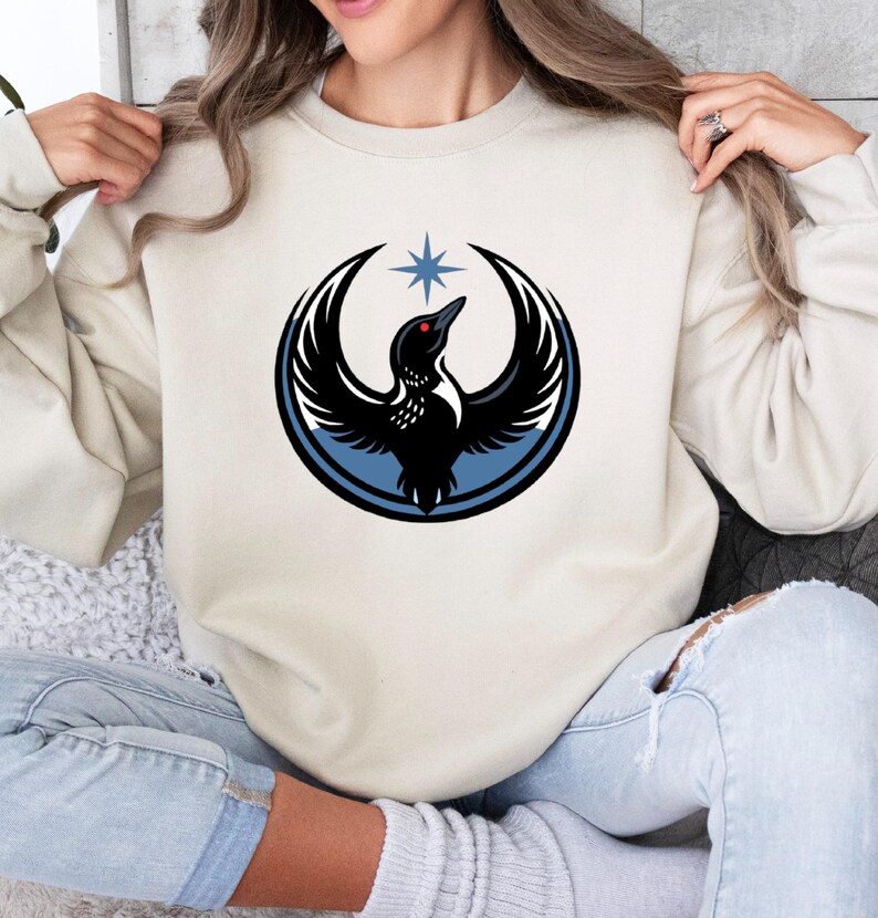 May include: Cream-colored sweatshirt featuring a black bird design within a blue circle, with a star above. The bird has a red eye and is set against a cream background. The sweatshirt is a casual style.
