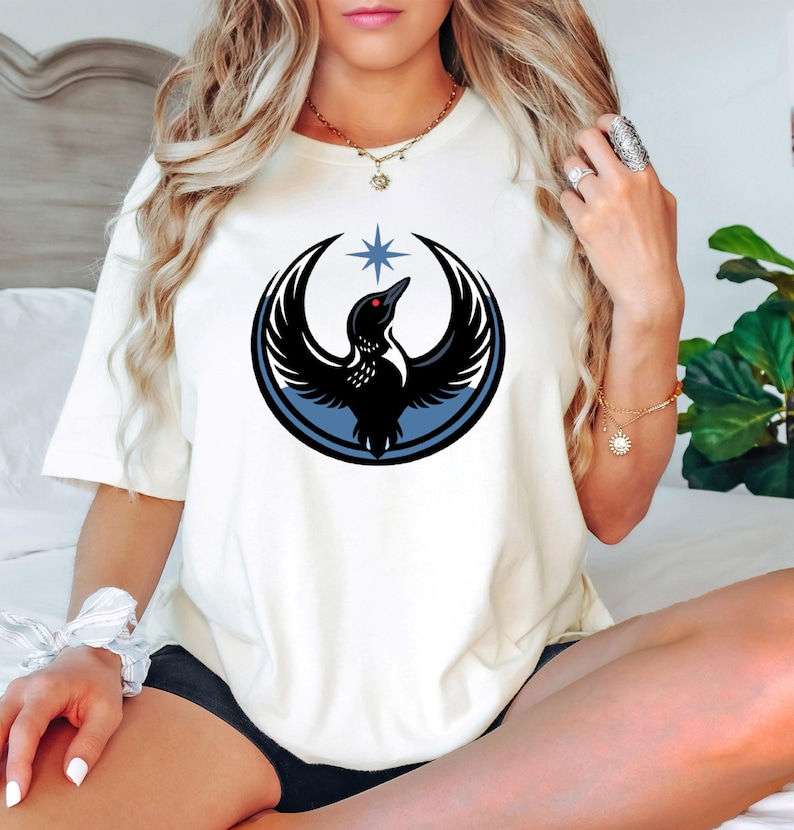 Puede incluir: Camiseta color crema con un dise&ntilde;o de p&aacute;jaro negro dentro de un c&iacute;rculo azul, con un ojo rojo y una estrella encima. El p&aacute;jaro tiene las alas extendidas. La camiseta es de un material suave.