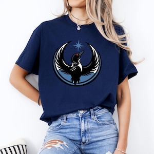 Puede incluir: Camiseta azul marino con un dise&ntilde;o de un ave con alas extendidas, una estrella y un dise&ntilde;o circular. El ave tiene un ojo rojo y est&aacute; sobre un fondo azul claro y blanco. Prenda de vestir informal.