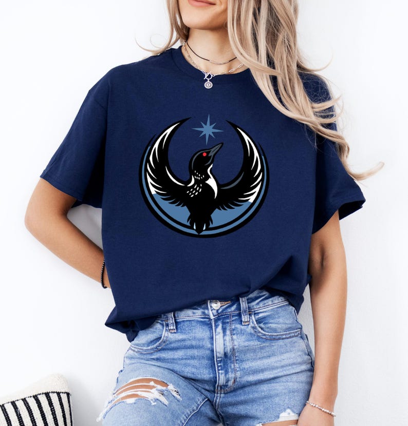 Puede incluir: Camiseta azul marino con un dise&ntilde;o de un ave con alas extendidas, una estrella y un dise&ntilde;o circular. El ave tiene un ojo rojo y est&aacute; sobre un fondo azul claro y blanco. Prenda de vestir informal.